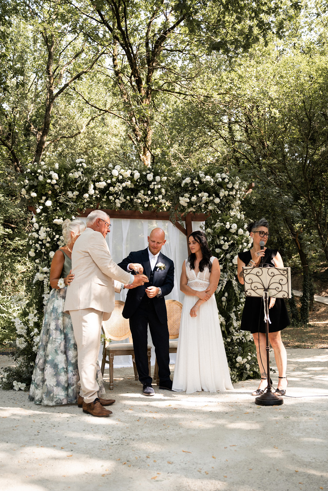 Les mariés et un proche pendant un rituel symbolique, entourés de l'arche fleurie, Sève officiant au micro à droite