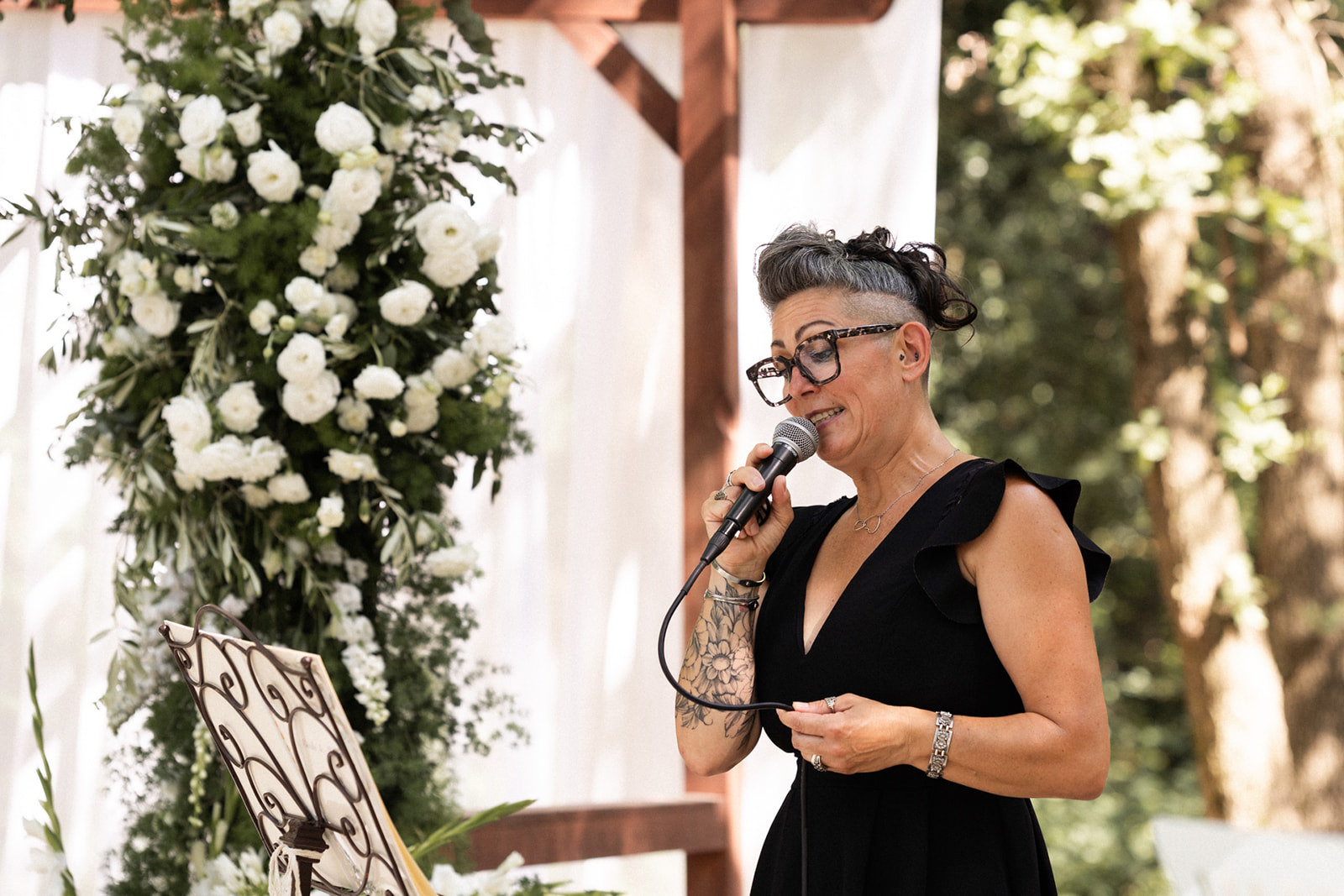 Sève, officiante, parle au micro devant l'arche fleurie blanche lors d'une cérémonie laïque en extérieur
